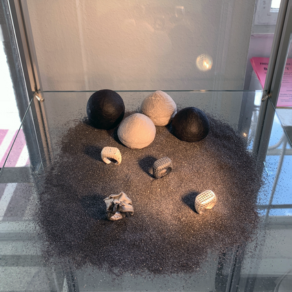 Ausstellungsdisplay mit Ringen, schwarzem Sand, grauen und schwarzen Steinen